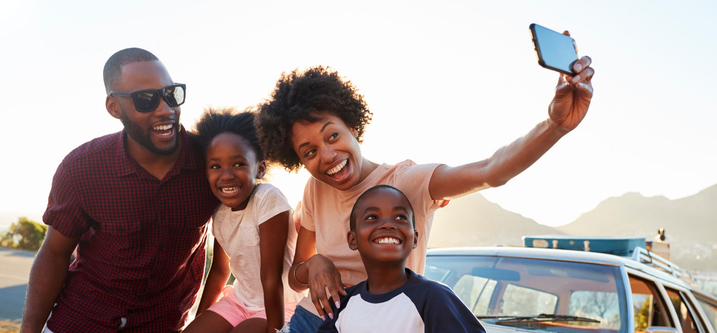 Image: Family posing for selfie next to car packed for road trip. (Photo Credit: Monkey Business / Adobe Stock)